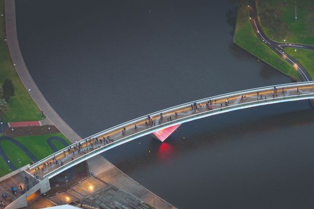 Aerial view of a broad curved pedestrian bridge spanning dark water, with many people walking along the illuminated deck. Curving paved paths, landscaped edges, and roadways are visible on both banks, while a red-lit structural element glows beneath the bridge. The image is a dusk or nighttime architectural infrastructure photograph.