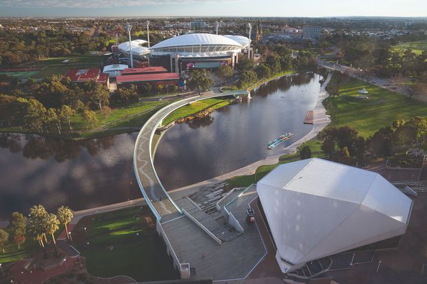 Aerial view of a landscaped urban precinct centered on a broad river or lake, with a sweeping pedestrian bridge, waterfront promenades, lawns, mature trees, and several large contemporary event or stadium buildings. The scene is photographed from above in warm daylight and emphasizes the civic architecture, water setting, and integrated public realm.