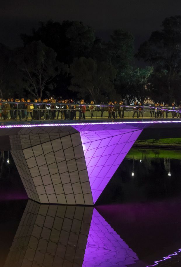Night-time photograph of a striking angular illuminated architectural structure spanning a dark body of water. The faceted, tiled or paneled form is lit vivid purple and reflected in the water, with a pedestrian bridge, decorative lights, and a crowd of people behind it among trees.