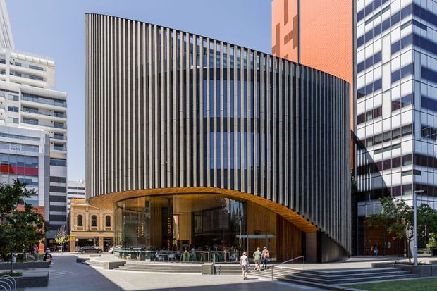 Street-level exterior view of a contemporary urban commercial building with a dramatic curved facade of closely spaced vertical fins, a sweeping timber-lined canopy, and a glazed recessed entrance. The building is surrounded by modern high-rise and heritage urban architecture, paved public space, and landscaping.