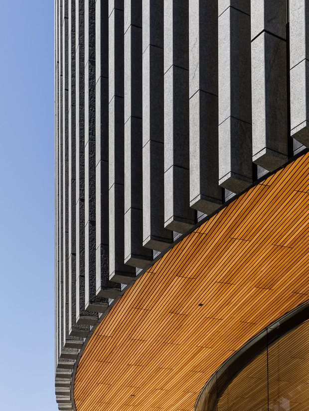 Close-up architectural photograph of a dramatic curved building facade. A warm orange timber-clad sweeping wall or soffit rises beneath repeated projecting dark grey vertical fins, creating a strong rhythm and contrast against the blue sky. The image focuses on the building envelope and geometric detailing, with no interior space visible.