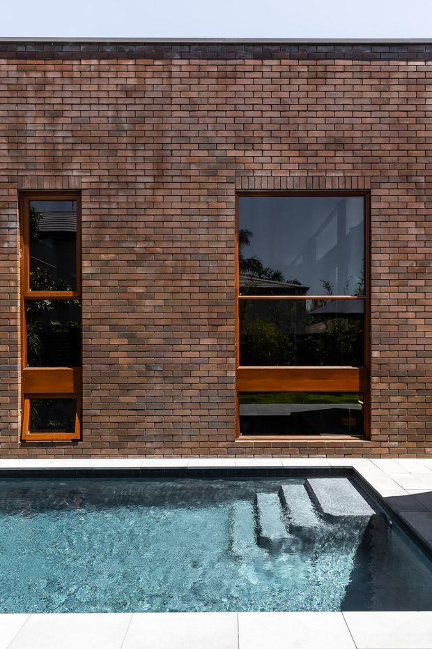 Exterior view of a contemporary brick building facade with narrow and large timber-framed windows overlooking a rectangular swimming pool. The warm brown brickwork and timber frames contrast with the dark reflective glazing, white pool coping, and blue water.
