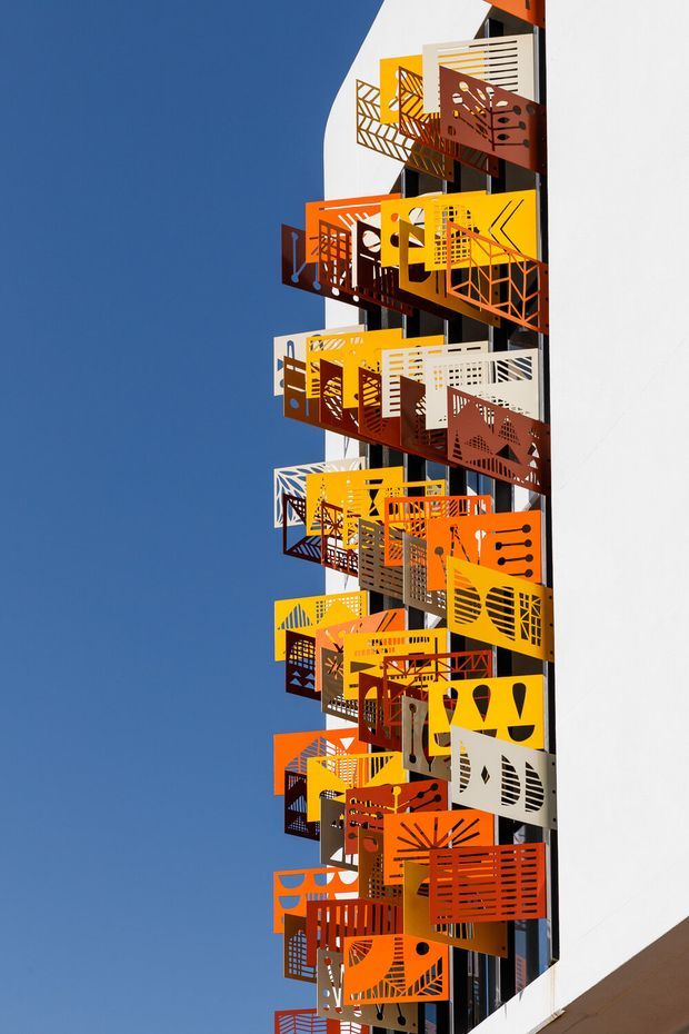 Close-up exterior view of a tall white building facade featuring a vertical stack of projecting metal balcony screens and privacy panels. The screens use bold geometric perforations and vivid yellow, orange, red, brown, white, and grey finishes, creating a highly graphic sculptural facade against a clear blue sky.