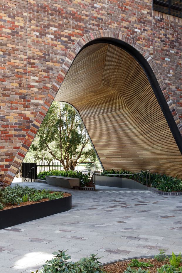 A landscaped architectural courtyard or public entry space framed by a dramatic arched brick structure. The arch contains a sweeping curved timber-lined soffit, with dark metal edging, integrated planting beds, paved surfaces, ramps, and seating. The design combines exposed brick masonry with warm timber detailing and contemporary landscape elements.