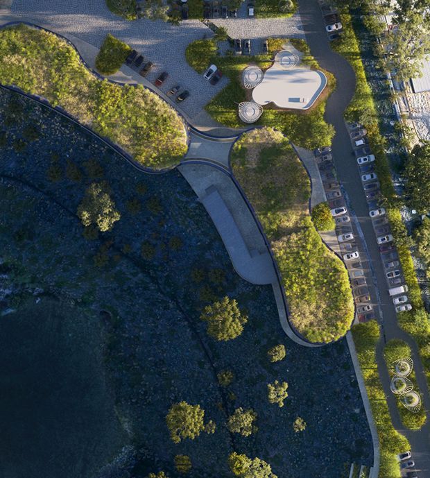 Aerial view of a landscaped public or commercial site beside a dark rocky waterfront or steep natural terrain. Curving paved driveways and pathways weave through planted green areas, with numerous parked cars along the edges. A distinctive light-coloured organic-shaped recreational or architectural feature sits near the upper right, accompanied by several circular sculptural elements. The site has a carefully designed, contemporary landscape layout.