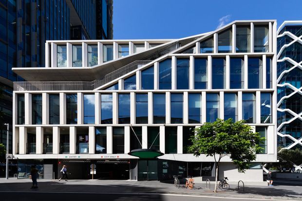 Street-level exterior view of a contemporary multi-storey commercial building in an urban setting. The facade combines extensive blue-tinted glazing with white structural framing, projecting terraces, a dramatic sloping roofline, and a prominent zigzag external structural feature on the adjacent section. Retail or commercial frontage occupies the ground level, with pedestrians and bicycles on the street.