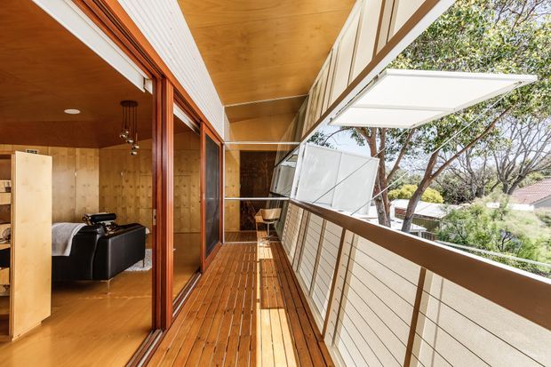 A bright elevated timber balcony or covered outdoor walkway running alongside a timber-clad interior room. The space features warm timber decking and ceiling boards, large sliding glazed doors with timber frames, a horizontal slatted privacy screen and a distinctive outward-opening awning window. A compact bedroom or living space with a black leather sofa is visible through the glazing.