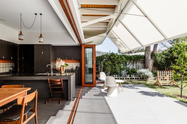 A bright contemporary indoor-outdoor entertaining area combining a dark timber kitchen with island bench, dining table and chairs, and a white-furnished covered terrace opening onto a landscaped garden. The glazed, retractable roof and exposed white structural framing create a strong connection between the kitchen and outdoor space, while patterned tile detailing and warm timber accents add character.