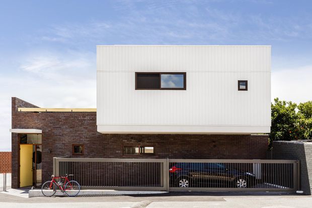 Street-facing view of a contemporary multi-level urban residence. A white vertically ribbed upper volume projects over a dark brick lower level, with narrow horizontal windows, a dark metal vehicle gate, and a recessed entry at the left. The composition has a clean, rectilinear modern facade with strong contrast between white cladding and brickwork.