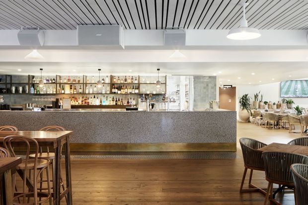 A bright contemporary hospitality interior centered on a long speckled stone bar with backlit liquor shelving, pendant lights, timber flooring, and café-style dining seating. The space also features a linear slatted ceiling, exposed services, indoor plants, and a television, creating a polished restaurant or hotel-bar atmosphere.