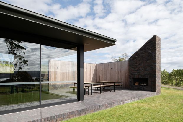 Contemporary single-storey house exterior with a broad flat roof, dark-framed floor-to-ceiling glazing, and an outdoor entertaining terrace. The terrace is enclosed by a vertical timber privacy fence and a striking freestanding triangular brick fireplace wall with built-in firewood storage and hearth. Brick paving extends around the outdoor dining area, with lawn and landscaped planting beyond.
