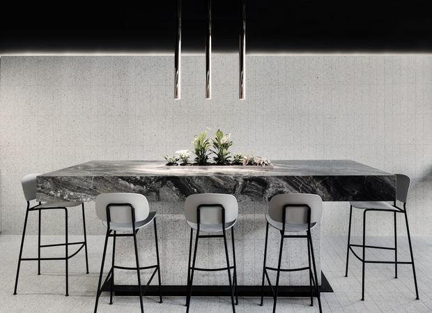 A refined contemporary dining area with a long dark veined stone island or dining table, surrounded by five black-framed bar stools with light upholstered seats. The space features pale textured wall and floor finishes, a dark ceiling, three slim pendant lights, and a small central floral arrangement. The composition is minimalist, monochromatic, and high-contrast.