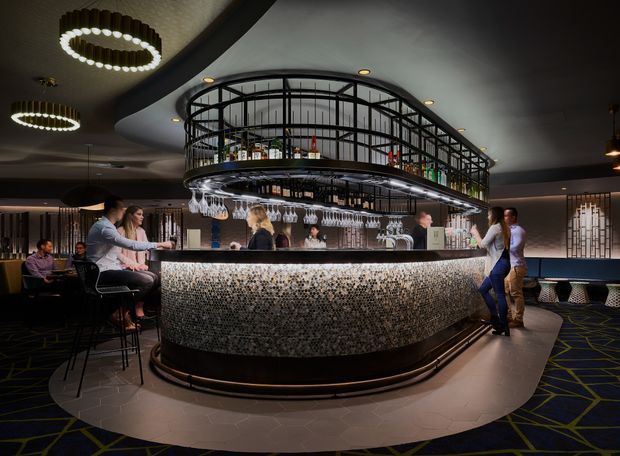 A sophisticated, dimly lit cocktail bar interior centered on a large curved island bar. The bar features a textured mosaic-style front, dark trim, illuminated shelving with bottles, suspended glassware, and a black metal overhead display structure. Guests sit on stools around the bar, with patterned carpet, dark walls, and decorative ring pendant lights contributing to a dramatic hospitality atmosphere.