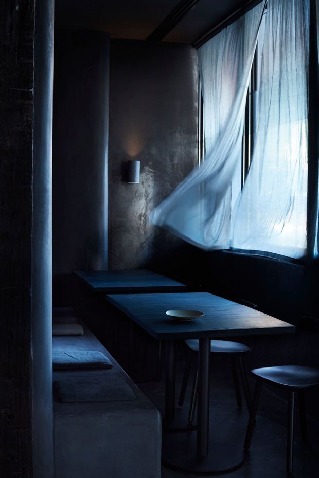 A dark, intimate dining area with built-in upholstered bench seating, simple dark timber tables, and low stools beside a large window. Sheer white curtains billow inward, creating a dramatic cool-blue glow against the deep charcoal walls and furniture. The space has a minimalist, moody hospitality-like atmosphere.