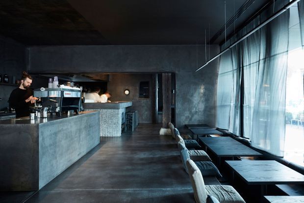 Dark, industrial-style café or hospitality interior with a concrete service counter and barista station on the left, a tiled counter face, polished dark flooring, exposed concrete walls and ceiling, and a long row of dining tables beside full-height glazed windows with sheer curtains. Black pendant fixtures and restrained furnishings create a moody, contemporary atmosphere.