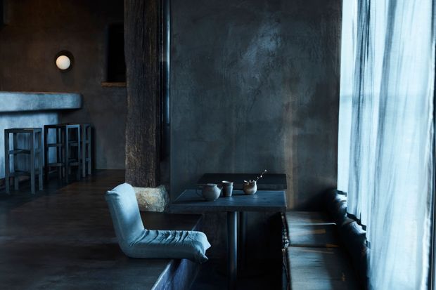 A moody, dark-toned interior showing a compact dining or lounge area beside floor-to-ceiling sheer curtains. A small dark table with ceramic vessels is paired with a sculptural upholstered chair and a long built-in or banquette-style bench. The space extends to a bar-height counter with stools on the left. Textured charcoal walls, dark flooring, a substantial timber column, and cool natural light create an atmospheric, minimalist setting.