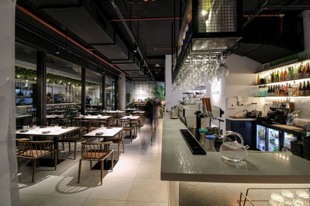 A contemporary restaurant or bar interior viewed from the service counter, with a large dining area to the left and a well-equipped bar to the right. The space features exposed black ceilings and services, dark-framed glazing, tiled flooring, timber dining tables and chairs, illuminated bottle shelving, hanging glassware, and polished hospitality counters. The atmosphere is sophisticated, industrial, and moody.