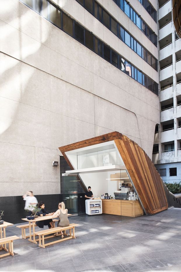 An urban outdoor café kiosk occupies a paved plaza between tall commercial buildings. The kiosk has a striking angular timber-clad canopy that folds down toward the ground, with a bright white service interior, timber cabinetry, counter equipment, and outdoor communal picnic-style seating. The surrounding architecture is tall, rectilinear, and concrete-panelled with bands of dark glazing and balconies. People are present around the seating and counter.