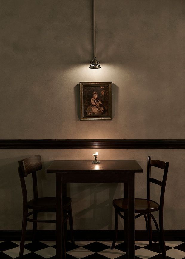 A moody, intimate dining nook with a dark wooden two-person table and matching traditional chairs against a textured taupe plaster wall. A small framed portrait is illuminated by a wall-mounted spotlight, while a candle sits on the table. Black-and-cream geometric checkerboard flooring and dark horizontal wall trim add a dramatic, historic character.
