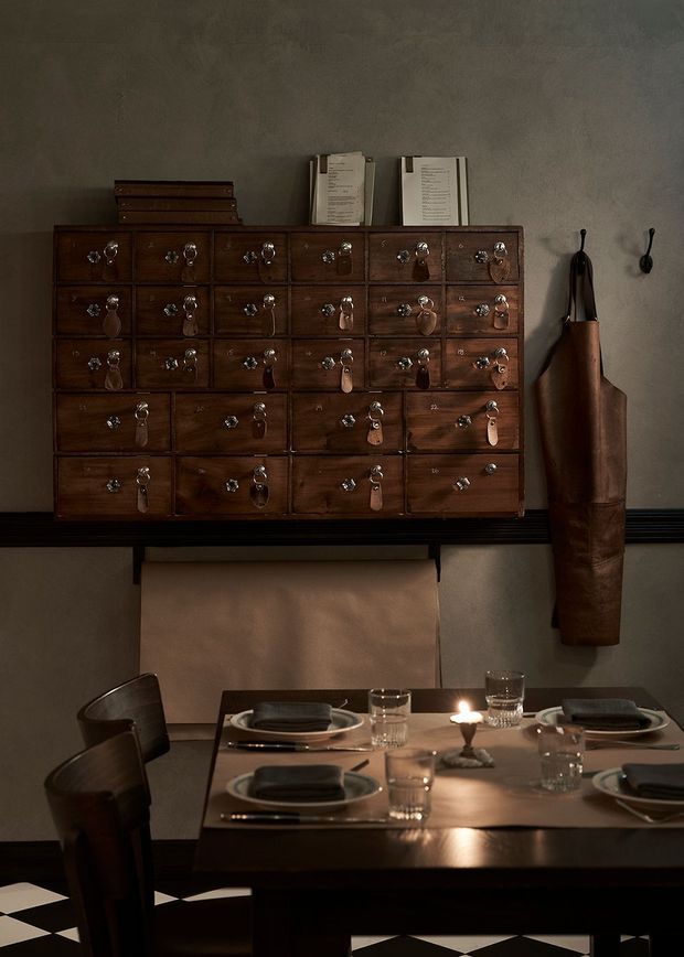 Moody, intimate dining area with a dark timber dining table set for four, leather-upholstered chairs, candlelight, and tableware. A large vintage-style wooden apothecary drawer cabinet with metal pulls is mounted on a textured grey wall, alongside a hanging leather apron. The room has a striking black-and-white checkered floor and warm, low lighting.