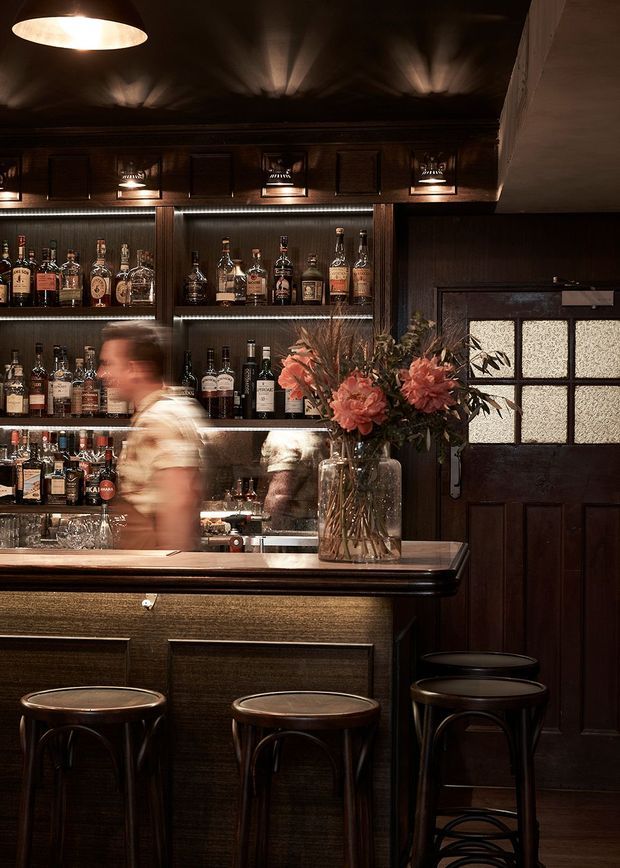 A moody, richly detailed bar interior with dark timber panelling, a wood-fronted counter, illuminated backbar shelving filled with bottles and glassware, pendant lighting, upholstered-free bar stools, and a dark timber entrance door with textured glass. A large floral arrangement adds a warm contrasting accent.