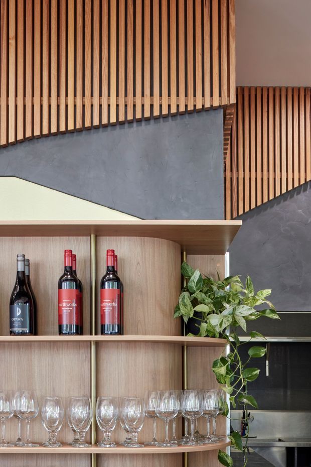 Close-up interior view of a refined hospitality-style bar or drinks cabinet. Curved light timber shelving displays wine bottles and rows of wine glasses, with a trailing green plant adding natural contrast. The background combines dark grey plaster or concrete-like wall surfaces with prominent vertical timber slat screening and concealed warm lighting. A glimpse of stainless-steel equipment is visible behind the shelving, suggesting a commercial bar or kitchen setting.