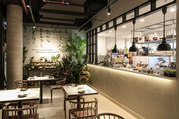 A warmly lit contemporary restaurant interior with dining tables and wooden chairs in the foreground, a long illuminated service counter and open commercial kitchen beyond, black-framed glazing, pendant lights, exposed services, tiled walls, concrete columns, and abundant indoor planting. A wall graphic provides a decorative focal point.