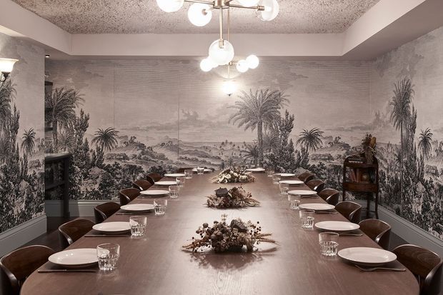 A formal dining room centred on a long wooden dining table with place settings for a large group. Dark upholstered dining chairs line both sides, while a dramatic monochrome pastoral mural covers the walls. Contemporary globe pendant lights hang from a textured ceiling, creating a refined, atmospheric entertaining space.