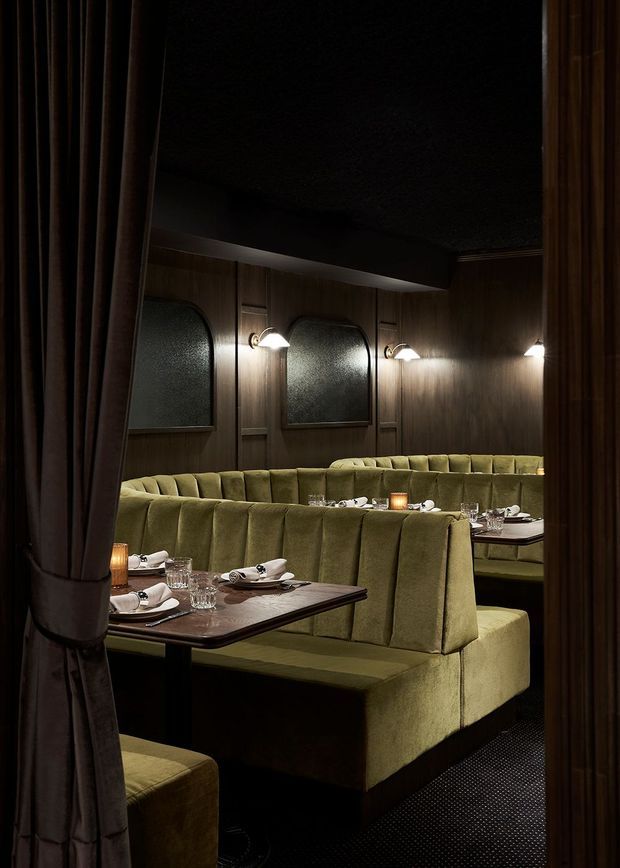A dimly lit, refined restaurant dining room viewed through heavy curtains. Olive-green upholstered booth seating surrounds dark timber dining tables set with white crockery, glassware and candles. The walls feature dark timber panelling, arched textured-glass panels and warm wall sconces, creating an intimate, theatrical atmosphere.