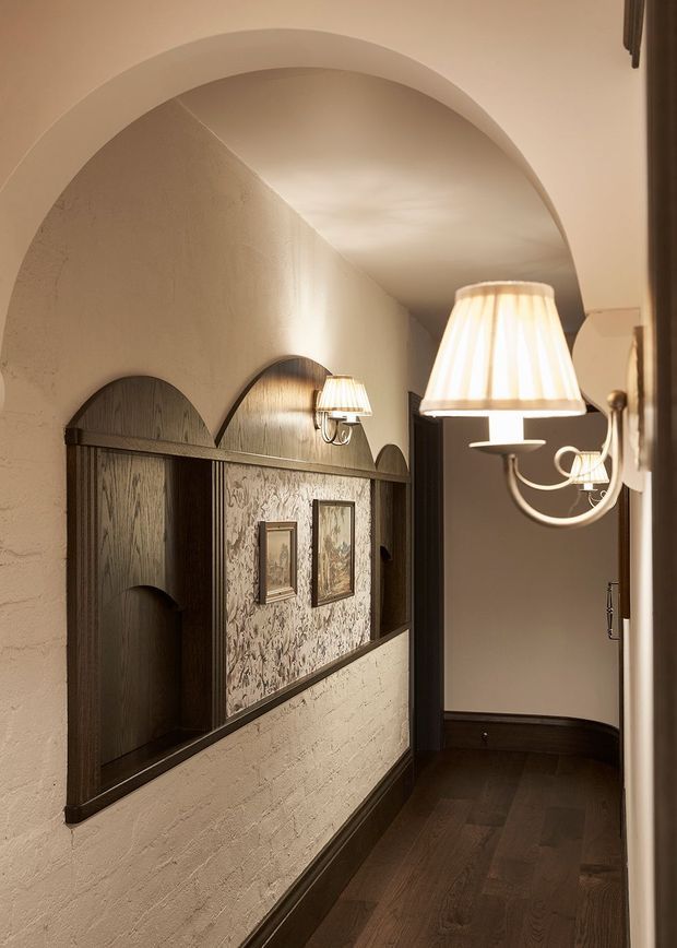 A warmly lit, narrow hallway featuring a large rounded plaster arch, textured cream walls, dark timber flooring and trim, and custom dark wood wall panelling with repeated arched niches. Decorative framed artwork and traditional wall sconces with pleated fabric shades add a refined, intimate character.