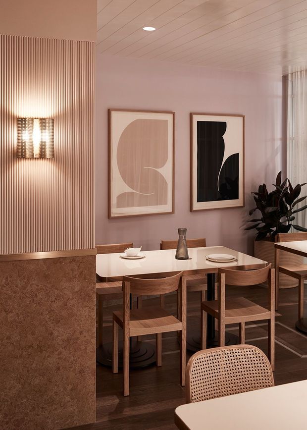 A refined dining area with a warm, contemporary interior. The room features a timber dining table and chairs, dark timber flooring, soft pink-beige walls, a vertically fluted wall with integrated lighting, framed abstract artwork, and a white slatted ceiling. A stone-clad partition and indoor plants add texture and contrast.