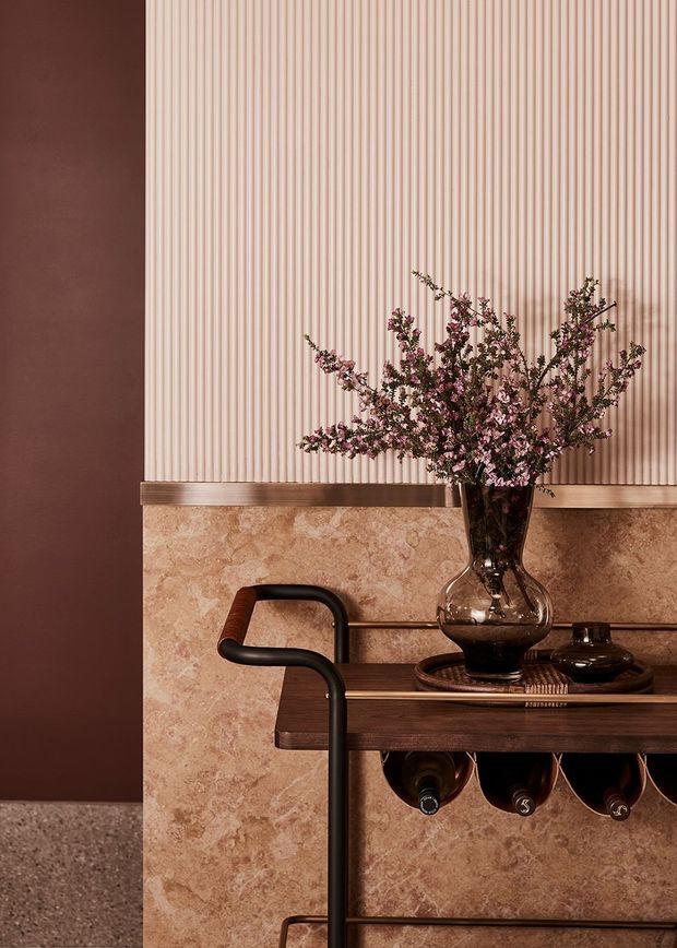 Close-up interior vignette featuring a dark wood and black metal bar or drinks trolley against a richly layered wall. The lower wall is clad in warm beige-pink natural stone, while the upper wall has narrow vertical fluted or reeded paneling in a pale blush tone. A smoked glass vase with pink flowering branches, a tray, and several wine bottles create a refined, warm decorative arrangement.
