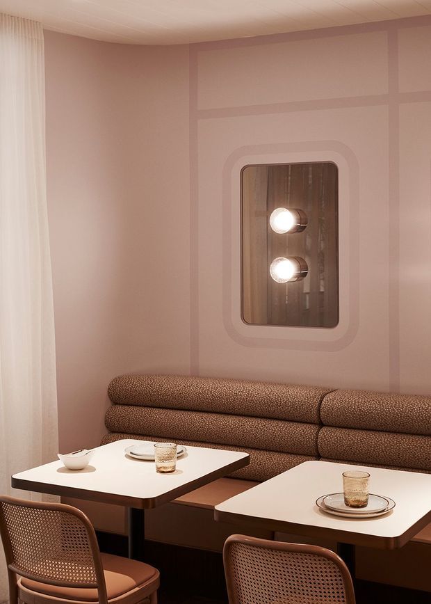 A warmly lit, refined dining nook or café-style seating area with blush-pink paneled walls, a built-in textured banquette, two small white tables, and woven cane dining chairs. Sheer curtains soften the light at the window, while a rounded rectangular wall mirror with integrated globe lighting forms a feature above the seating.
