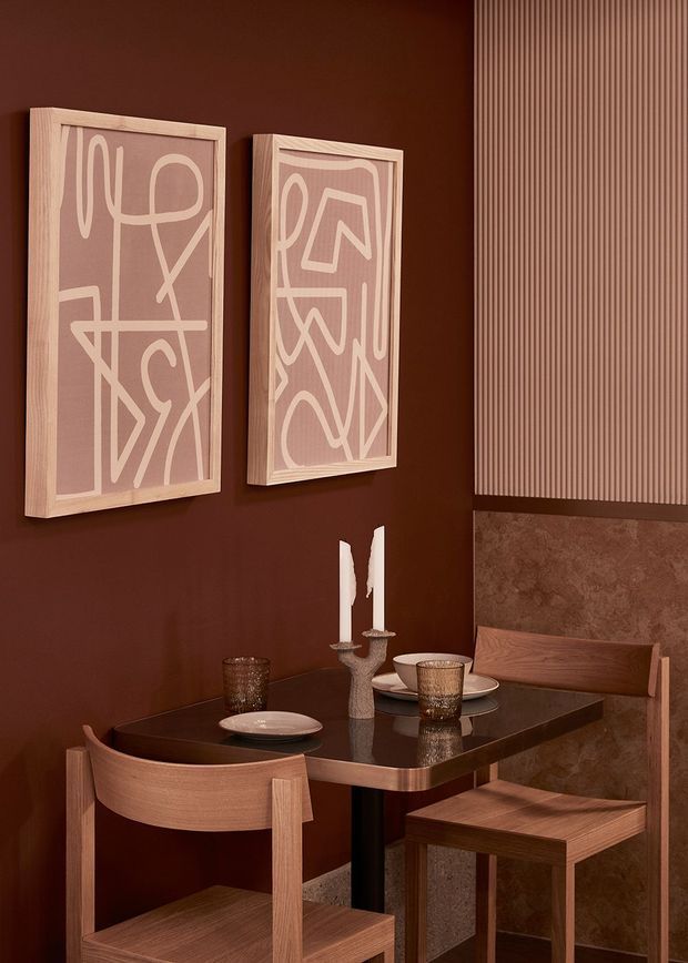A compact, richly coloured dining nook with a dark chocolate-brown wall, two framed abstract artworks, and a vertical fluted wall panel above a textured brown stone or plaster wainscot. A glossy dark dining table is set with ceramic plates, amber-toned glassware, candles, and sculptural tableware. Curved timber dining chairs and a simple timber stool add warmth, with a built-in upholstered bench along the right wall. The overall composition is intimate, refined, and contemporary with strong earthy tones and tactile surfaces.