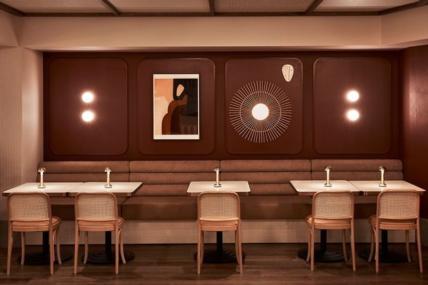 A sophisticated restaurant or hospitality dining space with a long upholstered banquette, individual marble-topped tables, cane-back timber chairs, warm integrated wall lighting, dark timber flooring, and richly toned brown wall panelling. Decorative artwork and a circular sunburst feature add an Art Deco-inspired character.