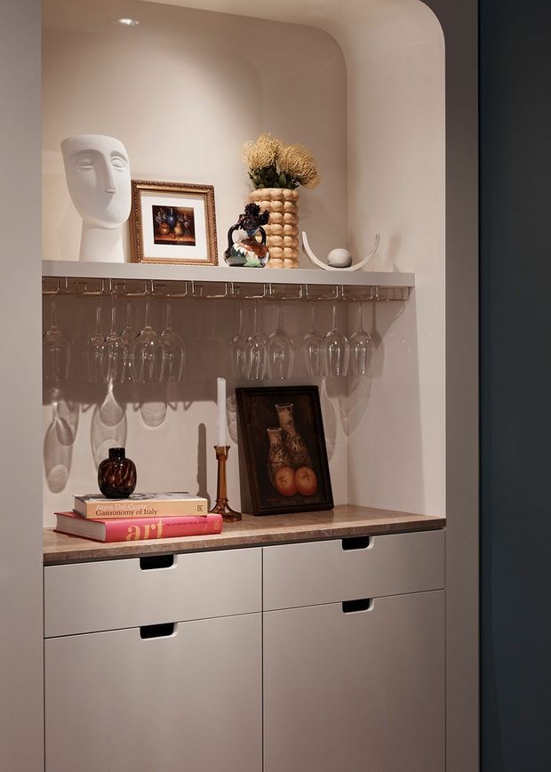 A compact built-in display and bar-storage niche with softly rounded plastered arch details. The upper shelf displays artwork, sculptural objects and dried flowers, while a hanging stemware rack holds wine glasses above a stone-look benchtop. Matte light cabinetry with integrated black pulls provides drawer and cupboard storage below. Warm ambient lighting creates a refined, curated atmosphere.