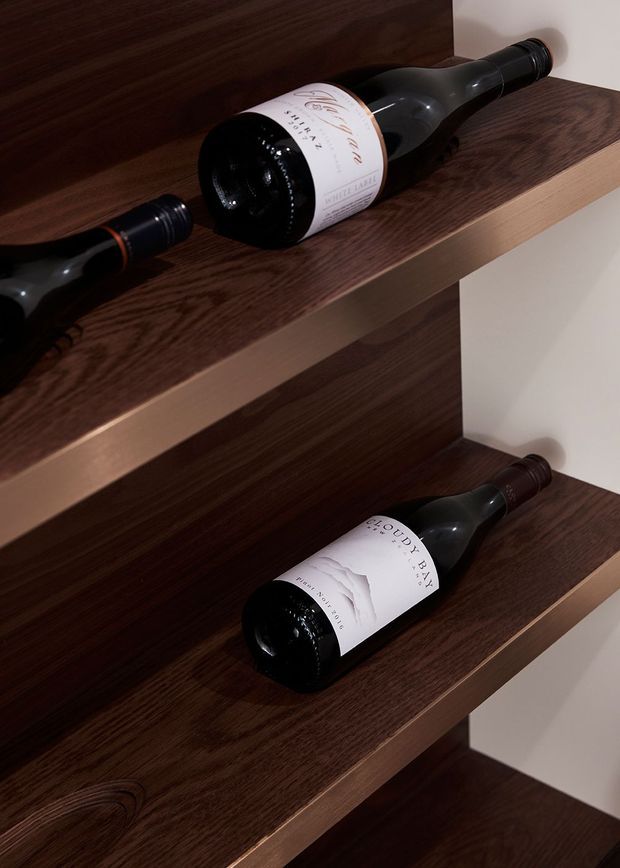 Close-up of dark timber wine-storage shelving with several wine bottles resting horizontally on individual shelves. The shelves have a rich dark wood grain and contrasting light-coloured wall backing, creating a refined built-in wine display.