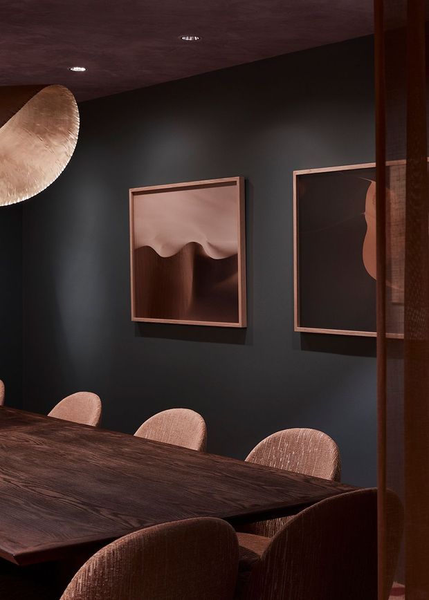 Moody, intimate dining room with a dark charcoal wall, richly stained timber dining table, upholstered dining chairs in warm brown and blush tones, and framed sculptural artwork. Soft, low-level lighting and a large textured pendant create a refined, dramatic atmosphere, with timber-framed glazing or screening at the right.