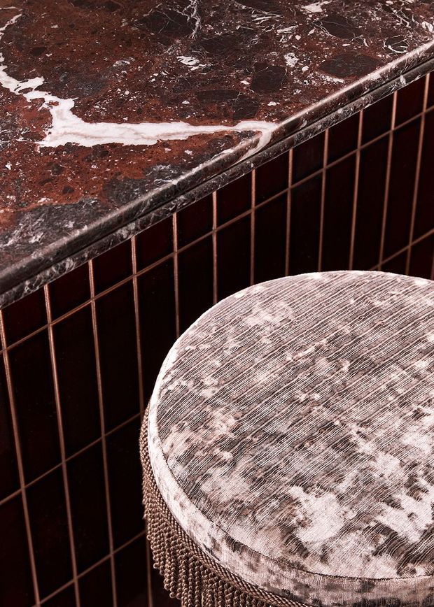 Close-up detail of a richly styled interior corner featuring a dark burgundy-brown marble or stone benchtop with prominent white veining, a dark reddish-brown tiled wall, and a round upholstered stool with a textured pale patterned seat and beaded trim. The image emphasizes luxurious materials, texture, and contrast.