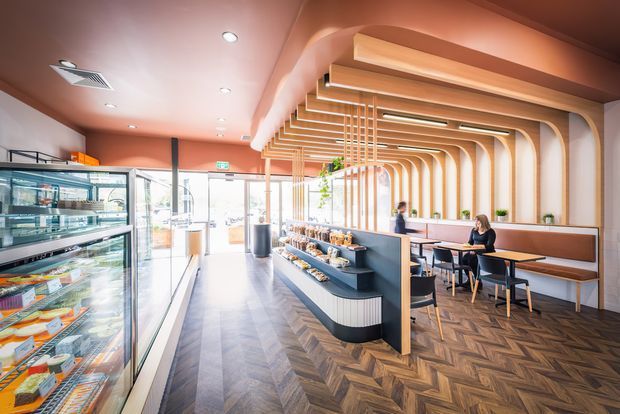 A bright contemporary café or bakery interior with refrigerated display cases along the left, a central food display island, and a compact dining area with banquette seating on the right. The space features an elaborate curved timber-look ceiling and wall framework, warm terracotta walls, pendant-style linear lighting, and a dark herringbone timber floor. Full-height glazing opens toward the street at the rear.