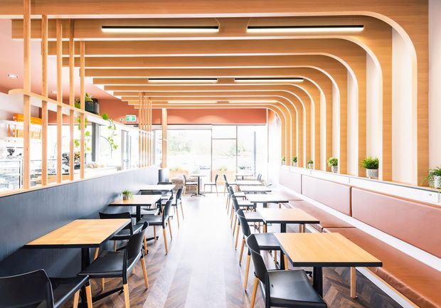 Bright contemporary commercial dining space with rows of timber tables and black chairs, a long banquette along the right wall, and a repeated curved timber ceiling and wall-frame structure. Full-height glazing, integrated linear lighting, indoor plants, and a herringbone timber floor create an airy, refined café or restaurant interior.