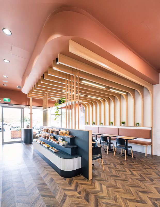 Bright contemporary café or casual dining interior with a curved timber slat ceiling and matching arched wall framework. A central dark service/display counter presents food, while upholstered banquette seating, small tables, and chairs line the glazed perimeter. The space has a warm blush-pink and timber palette, herringbone wood flooring, integrated linear lighting, and abundant daylight.