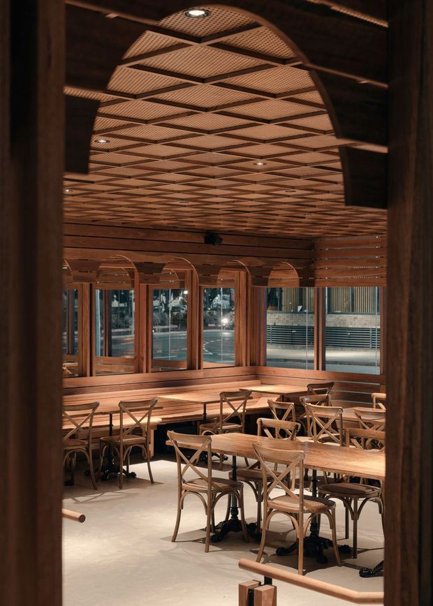 A warmly lit contemporary dining and hospitality interior viewed through a dark timber-framed opening. The room features extensive timber detailing, floor-to-ceiling windows, banquette seating along the perimeter, and multiple timber dining tables with cross-back chairs. A distinctive suspended geometric timber lattice ceiling with recessed downlights creates a strong architectural feature. Urban buildings are visible through the glazing.