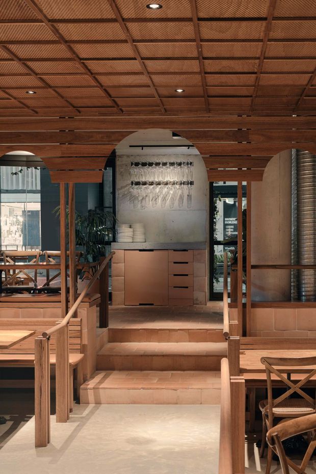 Warm, richly detailed hospitality-style interior with a central tiled stairway leading to a compact service or bar area. The space features extensive timber framing and joinery, a coffered perforated timber ceiling, terracotta-toned tiled steps and low walls, a concrete or plaster feature wall with hanging glassware, and seating areas visible on both sides. A large cylindrical metal duct forms a strong industrial element on the right.