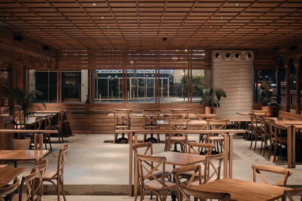 A warmly lit contemporary dining and hospitality interior with extensive timber detailing. The space features numerous timber dining tables and cross-back chairs, a raised timber banquette along the rear, large glazed openings, indoor plants, and a prominent timber-clad feature wall with circular ventilation or decorative openings. A gridded timber ceiling and dark timber wall framing create a layered, inviting restaurant or café atmosphere.