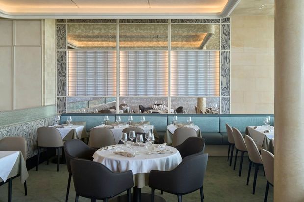 Elegant upscale restaurant dining room with multiple round tables dressed in white linens, upholstered dark and taupe dining chairs, and a long blue-green banquette. Decorative patterned wall panels frame mirrored or glazed sections, while horizontal blinds, pale stone walls, and a recessed illuminated ceiling create a refined contemporary interior.