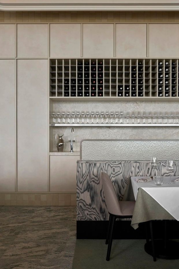 A refined dining and wine-storage area with full-height pale cabinetry, an open built-in wine rack, and shelves displaying rows of wine glasses. A textured stone or plaster backsplash frames a compact sink and tap. In the foreground, a dining table with a white cloth and upholstered seating sits beside a striking vertically grained timber feature partition. The palette is muted and warm, with pale beige, grey, and natural wood tones.