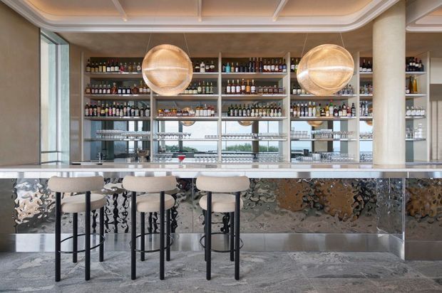 A refined bar interior with a long metallic-faced counter, four upholstered black-frame bar stools, extensive back-bar shelving filled with bottles and glassware, mirrored panels, and two large spherical pendant lights. The space features a pale stone floor, warm neutral finishes, curved architectural detailing, and views through glazing behind the bar.