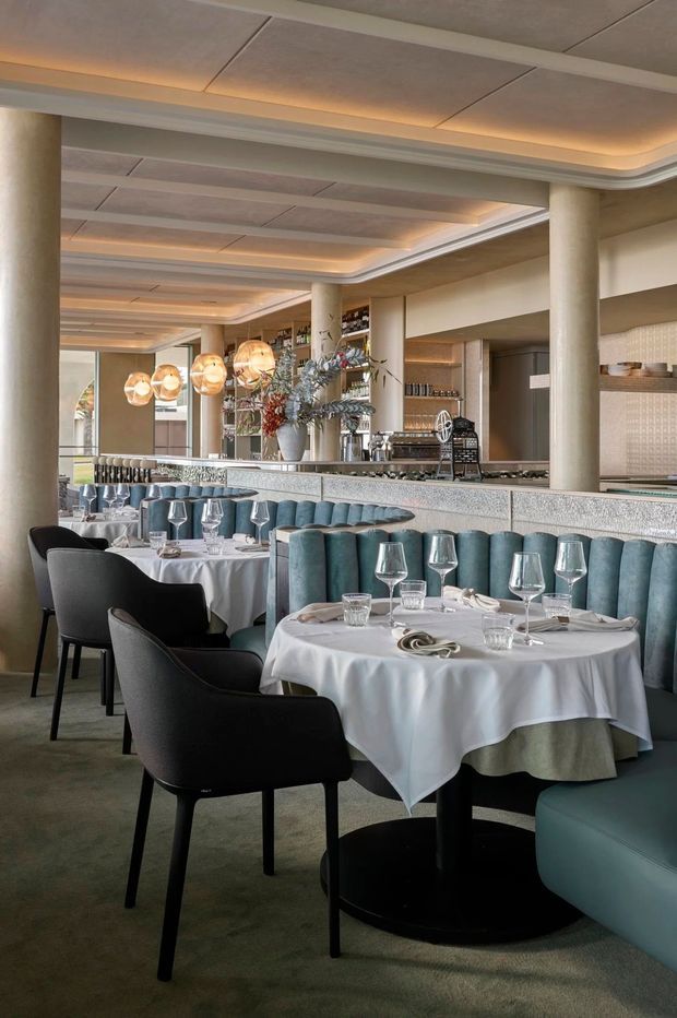 A refined restaurant dining room with multiple round, white-clothed tables set with wine glasses and tableware. Curved teal upholstered banquettes and black dining chairs sit on a muted carpet, while a bar and open service counter run along the rear. Large cylindrical columns, layered coffered ceiling panels, concealed warm lighting, decorative pendant lights, and light textured finishes create an elegant contemporary hospitality interior.