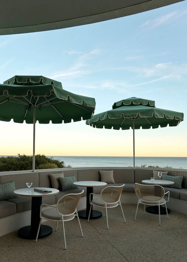 A refined outdoor terrace overlooking the ocean, furnished with curved upholstered banquette seating, small round café tables, and woven lounge chairs. Large dark green scalloped umbrellas provide shade, while a curved overhead canopy frames the open coastal view. The setting has a calm, upscale hospitality feel with a muted neutral and green palette.